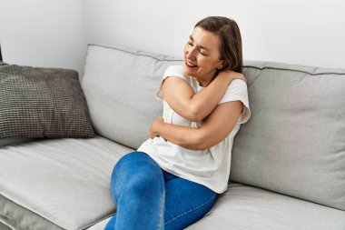 Middle age hispanic woman smiling sitting on the sofa hugging herself at home