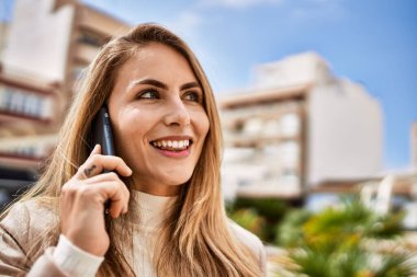 Genç sarışın kadın gülümsüyor. Kendine güveni tam. Akıllı telefondan konuşuyor.