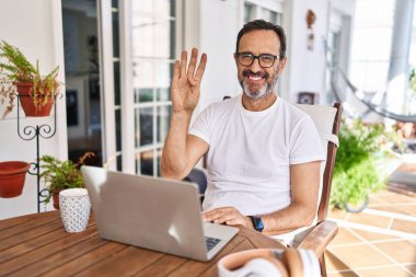 Orta yaşlı bir adam evde bilgisayar kullanıyor. Dört numarayı gösteriyor ve işaret ediyor. Kendinden emin ve mutlu bir şekilde gülümsüyor.. 