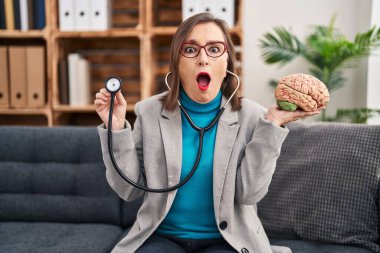 Middle age hispanic woman working at therapy office holding brain afraid and shocked with surprise and amazed expression, fear and excited face. 