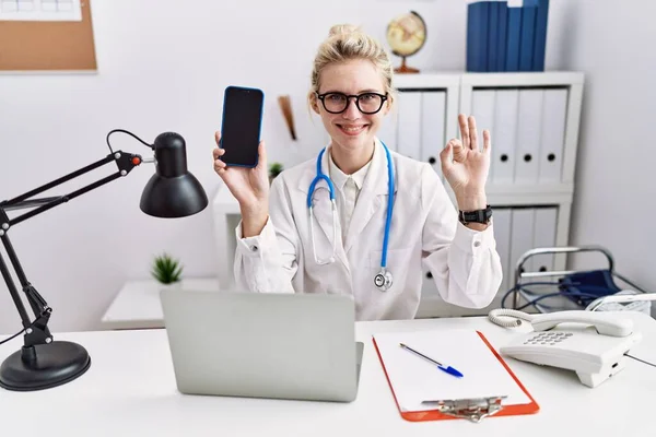 Young Doctor Woman Holding Smartphone Showing Blank Screen Doing Sign — Foto de Stock