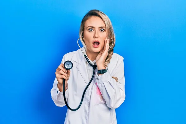 Beautiful young blonde doctor woman holding stethoscope afraid and shocked, surprise and amazed expression with hands on face