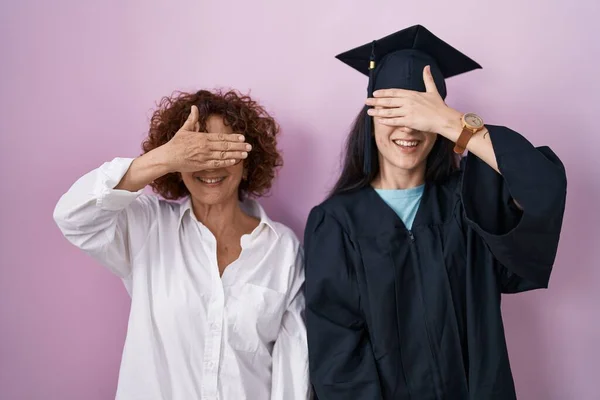 卒業キャップと式のローブを身に着けているヒスパニック系の母親と娘は笑顔で 驚きのために目をカバー顔に手で笑っています 視覚障害の概念 — ストック写真