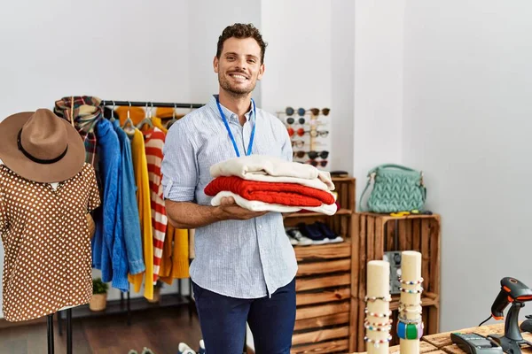 Young hispanic shopkeeper man smiling happy holding stack of sweater at clothing store.