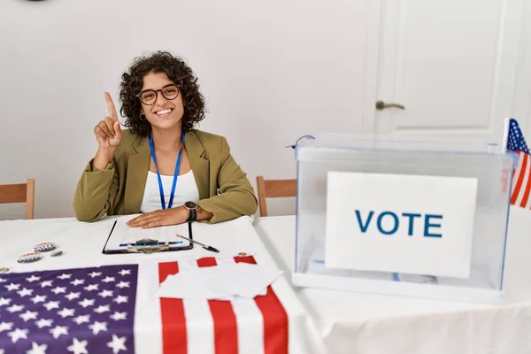 Young hispanic woman at political election sitting by ballot showing and pointing up with finger number one while smiling confident and happy.