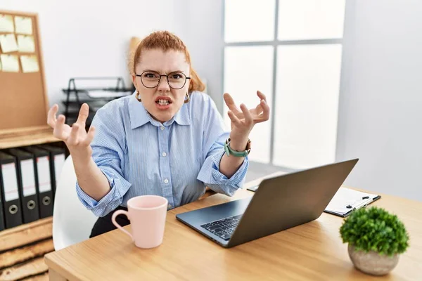 Young redhead woman working at the office using computer laptop shouting frustrated with rage, hands trying to strangle, yelling mad