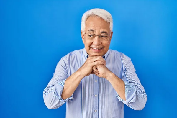 Hispanic senior man wearing glasses laughing nervous and excited with hands on chin looking to the side