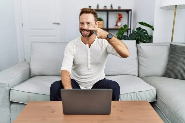 Middle age man using laptop at home pointing with hand finger to face and nose, smiling cheerful. beauty concept