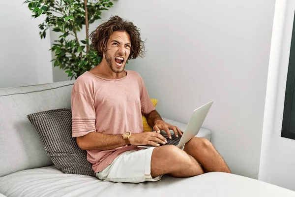 Young hispanic man sitting on the sofa at home using laptop angry and mad screaming frustrated and furious, shouting with anger. rage and aggressive concept.