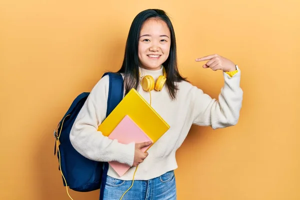 Young chinese girl holding student backpack and books looking confident with smile on face, pointing oneself with fingers proud and happy.