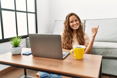 Güzel İspanyol kadın evde bilgisayar kullanıyor. Gülümsüyor, mutlu bir şekilde elini ve parmağını yana doğru gösteriyor. 