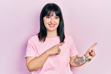 Gündelik kıyafetler giyen, gülümseyen ve kameraya bakan genç İspanyol kadın iki eliyle ve parmaklarıyla kenarı işaret ediyor.. 