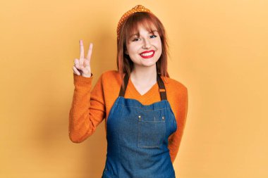 Kızıl saçlı, önlük takan, gülümseyen ve kameraya göz kırpan genç bir kadın zafer işareti yapıyor. İki numara.. 