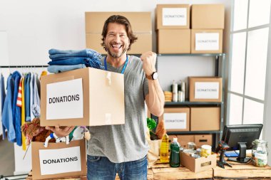 Gönüllü olarak bağış kutusunu tutan yakışıklı orta yaşlı bir adam mutlu ve heyecanlı bir şekilde kollarını kaldırıp, gülümseyip başarı için haykırıyor. kutlama konsepti. 