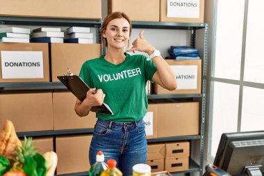 Bağışta gönüllü tişört giyen genç esmer kadın telefonda konuşurken gülümseyerek el ve parmaklarıyla telefon hareketi yapıyor. iletişim kavramları. 