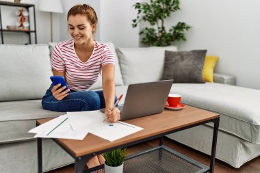 Akıllı telefon ve dizüstü bilgisayar kullanan genç bir kadın evdeki kanepede oturuyor.