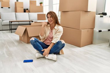 Orta Doğulu yaşlı bir kadın, yeni evinde yerde oturmuş gülümsüyor, gülümsüyor ve başparmağını kaldırarak yüzünü işaret ediyor.. 