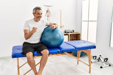 Orta yaşlı İspanyol bir adam ağrı kesici kliniğinde elinde pilates topu tutarak kamerayı mutlu ve komik yüzüyle işaret ediyor. iyi enerji ve titreşimler. 