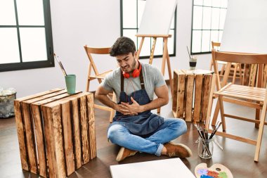 Young hispanic man sitting at art studio with hand on stomach because indigestion, painful illness feeling unwell. ache concept. 