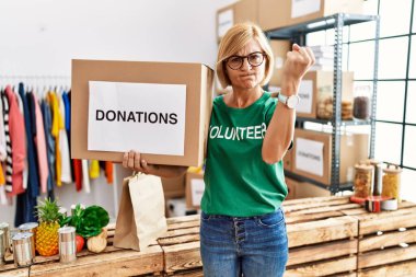 Middle age blonde woman wearing volunteer t shirt holding donations box annoyed and frustrated shouting with anger, yelling crazy with anger and hand raised 