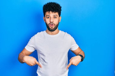 Sakallı, beyaz tişörtlü genç bir Arap. İlan veren parmaklarıyla işaret ediyor. Şaşırmış yüzü ve ağzı açık. 