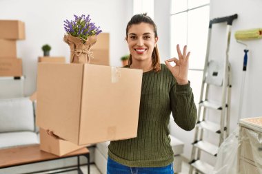 Genç İspanyol kadın hareket halindeyken karton kutu tutuyor tamam işareti yapıyor parmaklarıyla, gülümseyerek mükemmel bir sembol oluşturuyor. 