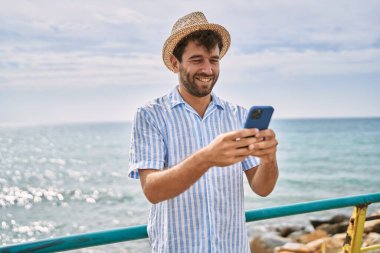Genç İspanyol adam sahilde akıllı telefon kullanarak mutlu bir şekilde gülümsüyor.
