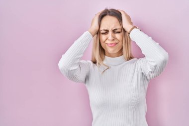 Pembe arka planda beyaz süveter giyen genç sarışın kadın baş ağrısı yüzünden çaresiz ve stresli çünkü ağrı ve migren. Eller başının üstünde.. 