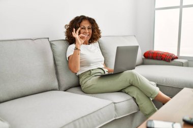 Orta yaşlı İspanyol kadın evde bilgisayar kullanıyor parmaklarıyla imza atıyor, gülümsüyor ve mükemmel bir sembol oluşturuyor. 