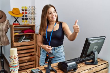 Perakende dükkanında açık mesajla pankart tutan genç esmer kadın gururlu görünüyor, gülümsüyor yan tarafa baş parmak işareti yapıyor. 