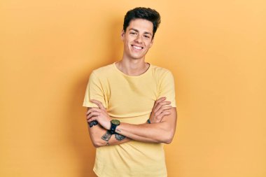 Sarı günlük tişört giyen genç İspanyol adam kollarını kavuşturup kameraya bakarken gülümsüyor. pozitif kişi. 