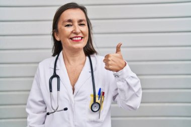 Middle age hispanic woman wearing doctor uniform and stethoscope smiling happy and positive, thumb up doing excellent and approval sign 