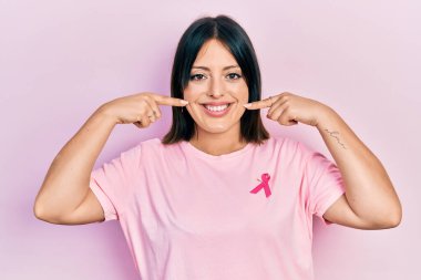 Gömleğine pembe kanser kurdelesi takmış neşeli gülümseyerek parmakları ve ağzıyla işaret eden genç İspanyol kadın. diş sağlığı kavramı. 