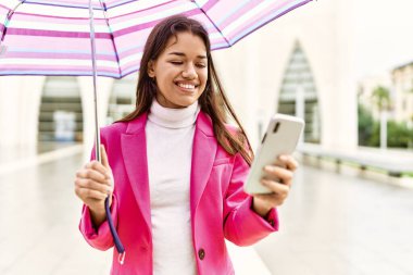 Young latin woman using smartphone holding umbrella at street