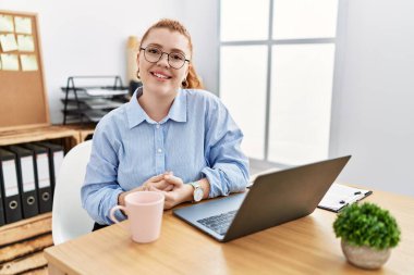 Ofiste çalışan genç kızıl saçlı kadın bilgisayarlı dizüstü bilgisayar kullanıyor ve parmaklarını çapraz yapıp gevşek ve neşeli gülümsüyor. Başarı ve iyimserlik 