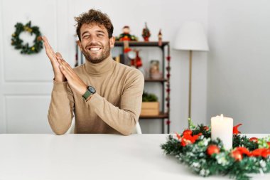 Masada sakallı genç yakışıklı adam Noel süslemeleri ile oturuyor ve alkışlıyor ve mutlu, neşeli, gururlu elleri birleştiriyor. 