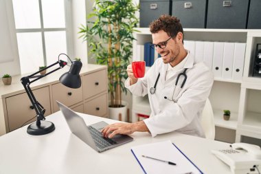 Doktor üniforması giyen, kahve içen genç İspanyol bir adam klinikte video görüşmesi yapıyor.