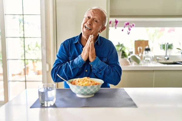 Senior man with grey hair eating pasta spaghetti at home praying with hands together asking for forgiveness smiling confident.