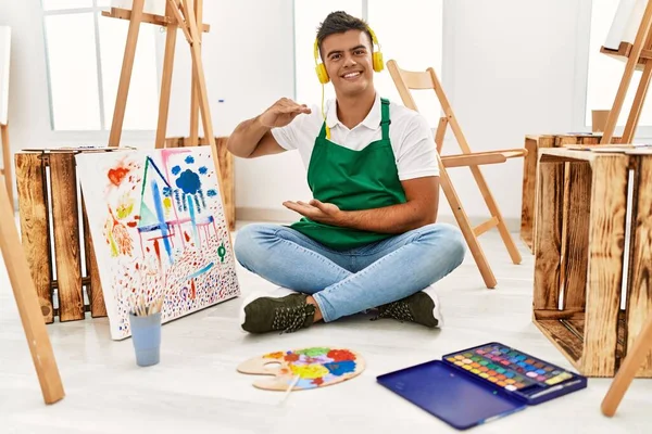 Young hispanic man at art studio gesturing with hands showing big and large size sign, measure symbol. smiling looking at the camera. measuring concept.