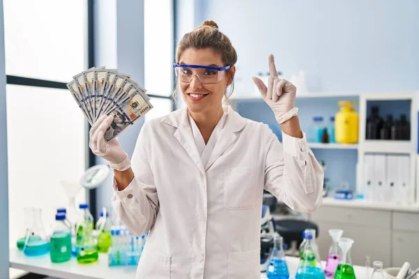 Young woman working at scientist laboratory holding money surprised with an idea or question pointing finger with happy face, number one