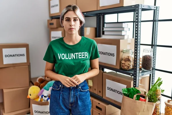 Young beautiful woman wearing volunteer t shirt at donations stand with serious expression on face. simple and natural looking at the camera.
