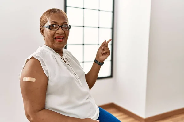 Mature hispanic woman getting vaccine showing arm with band aid with a big smile on face, pointing with hand finger to the side looking at the camera.