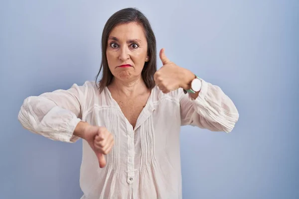 Middle age hispanic woman standing over blue background doing thumbs up and down, disagreement and agreement expression. crazy conflict