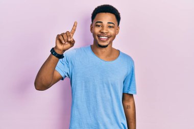 Genç Afro-Amerikan adam günlük mavi tişört giyer ve bir numaralı parmağını göstererek gülümserken kendinden emin ve mutlu. 