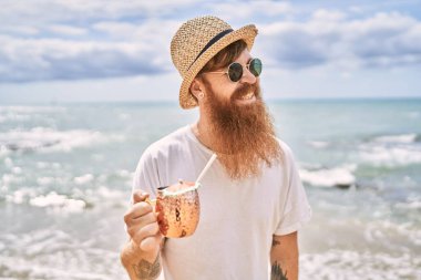 Genç kızıl saçlı adam gülümsüyor sahilde kokteyl içiyor..