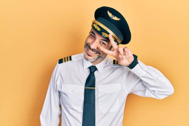 Handsome middle age man with grey hair wearing airplane pilot uniform doing peace symbol with fingers over face, smiling cheerful showing victory 