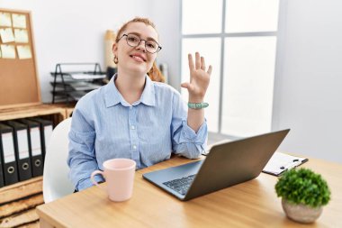 Ofiste çalışan genç kızıl saçlı kadın bilgisayarını kullanarak beş numaralı parmaklarıyla işaret ederken kendinden emin ve mutlu bir şekilde gülümsüyor.. 