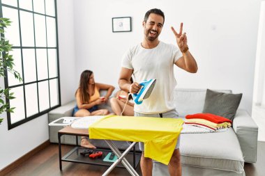 Genç İspanyol adam evde ütü yapıyor, gülümsüyor kameraya göz kırpıyor parmaklarıyla zafer işareti yapıyor. İki numara.. 