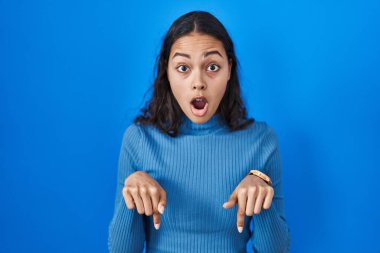 Genç Brezilyalı bir kadın, mavi arka planda dikilip parmaklarını göstererek reklamını yapıyor, şaşırmış yüzünü ve ağzını açıyor. 