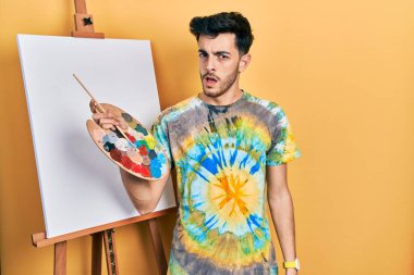 İspanyol asıllı genç adam paletle resim çiziyor. Şok olmuş yüz ifadesiyle duruyor. Şüpheci ve alaycı bakıyor. 